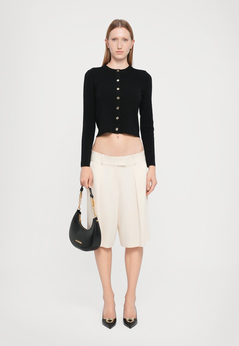 Black cropped cardigan with gold buttons, cream pleated shorts, black handbag with chain handle, and pointed toe black heels.