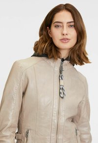 Lichtbeige leren jack met een gladde textuur, ritssluiting en zwarte capuchon. Heeft decoratieve trekkoorden en zijzakken.