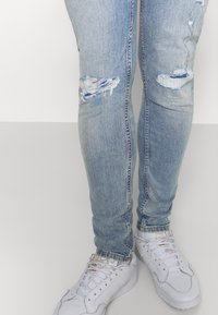 Persona vestida con jeans skinny desgastados de color azul claro y zapatillas Adidas blancas, de pie contra un fondo blanco liso.