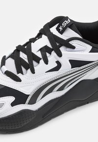 Svart och vit atletisk sneaker med en blandning av texturerade material, framträdande snören, smala silverdetaljer och en tjock, mönstrad sula.