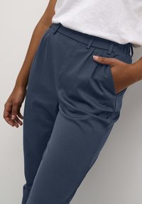 Personne portant un pantalon bleu marine taille haute avec une ceinture élastique et une chemise blanche rentrée, la main dans la poche, contre un fond simple.