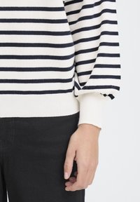 Pull en maille à rayures horizontales bleu marine et blanches, avec des poignets côtelés et une texture douce. Associé à un pantalon en tissu foncé.