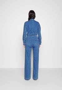 Blå denimoverall med en figursydd bodice, långa ärmar, vida byxor och subtila sömnadsdetaljer. Bakifrån syns två bakfickor.