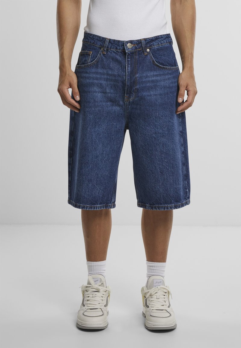 Homme portant un short en denim bleu foncé jusqu'aux genoux, des chaussettes blanches, des baskets blanches et un débardeur blanc sur un fond uni.