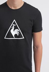 Svart bomull t-shirt med en vit triangulär logotyp av en tupp i en stiliserad design, korta ärmar och en figurnära siluett.