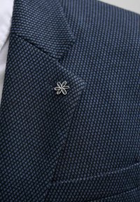 Tumedat sinise mustriga bleiser, millel on väike lilleline krae pannal, valmistatud tekstureeritud kangast geomeetrilise disainiga ja struktureeritud kraega.