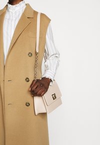 Beige Wollweste mit doppelreihigen Knöpfen, kombiniert mit einer weiß gestreiften Bluse. Hellrosa Handtasche mit goldener Ausstattung und Kettenriemen.