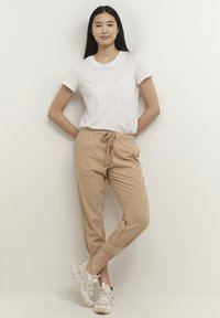 Bež lahke obrezane hlače z vrvico v pasu, združene z oprijetim belim majico in masivnimi belimi supergami na nevtralnem ozadju.