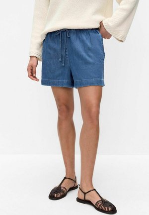 Personne portant un short en jean bleu avec cordon, un pull en maille crème et des sandales plates tressées noires, debout devant un fond blanc uni.