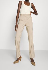Beige pinstripede højtaljede bukser parret med en tætsiddende let beige ribbet tanktop. Modellen bærer guldfarvede sko i slangeskindsmønster.
