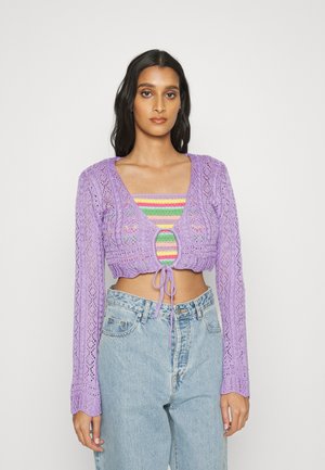Cardigan roxoa cropped com mangas longas e padrão de tricô de renda. Amarrado na frente com um cordão, sobreposto a uma blusa multicolorida.