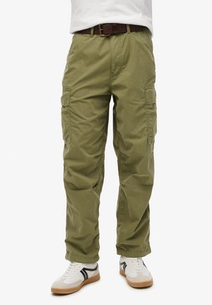 Olijfgroene cargobroek met twee zijzakken, relaxte pasvorm en een tailleband met een bruine riem. Draagt met witte sneakers en zwarte accenten.