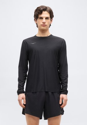 Νεαρός άνδρας με μαύρο αθλητικό μπλουζάκι μακρυμάνικο και αντίστοιχο σορτς, στέκεται μπροστά σε ένα απλό ανοιχτό γκρι φόντο.