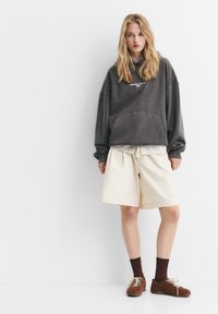 Übergroßer dunkelgrauer Sweatshirt mit einer vorderen Tasche, kombiniert mit cremefarbenen Shorts. Braune Wildlederschuhe und burgunderfarbene Socken runden das Outfit ab.