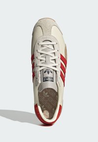 Beige leather sneaker with red suede accents, three red stripes, and a textured rubber sole. Rounded toe and lace-up design.