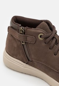 Hnědé semišové kotníkové boty s bočním zipem, polstrovaným límcem a texturovanou síťovinou. Podešev je světle béžová s hladkým povrchem.