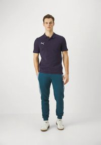 Tyrkysová polokošeľa s bielym logom Puma, skombinovaná s tyrkysovými teplákmi s bočnými akcentmi a bielymi teniskami. Štruktúrovaný strih, neformálny štýl.