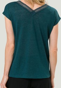 Haut à manches courtes teal en tissu léger. Présente un col en V avec des détails tissés le long de l'encolure arrière. Texture lisse.