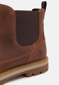Bottines en cuir marron avec des côtés élastiques, semelle en caoutchouc et semelle intermédiaire beige contrastante. Présente un logo embossé sur le côté extérieur.