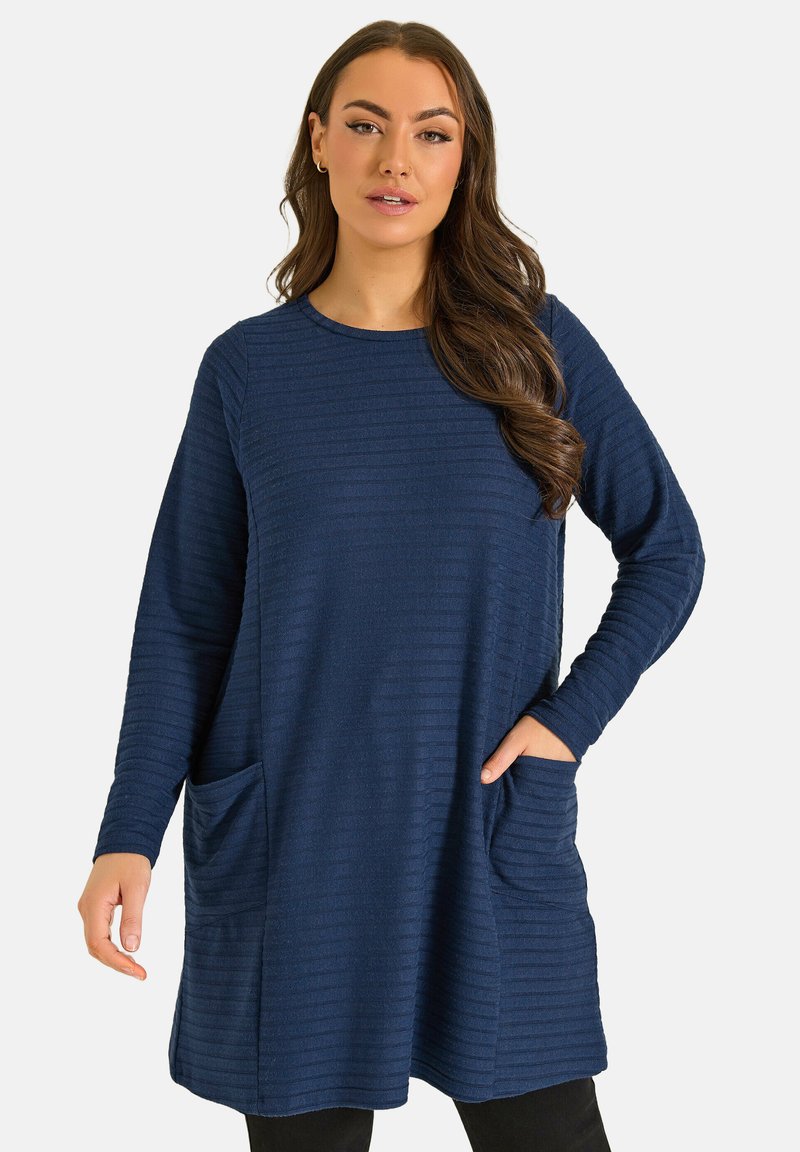 Donna con capelli castani lunghi che indossa una tunica blu navy a maniche lunghe con una texture e due tasche frontali, in piedi su uno sfondo semplice.