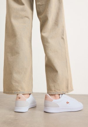 White sneakers with a smooth leather finish, featuring a beige suede accent on the heel and subtle branding. Paired with light beige pants.