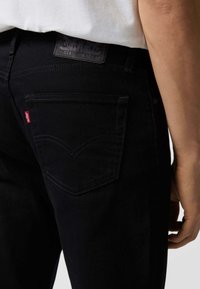 Vaqueros de mezclilla negra con un diseño ajustado. Incluyen un bolsillo trasero con una etiqueta roja de Levi's y una etiqueta de cuero en la cintura.