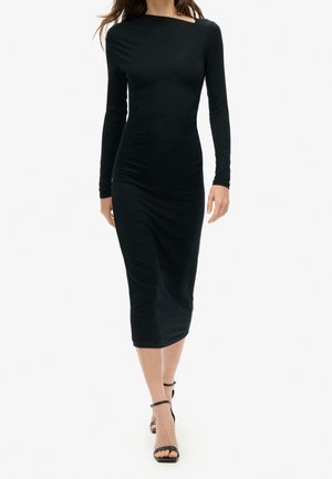 Kobieta idąca w dopasowanej, czarnej sukience midi z długim rękawem i dekoltem w łódkę oraz w czarnych sandałkach na wysokim obcasie z paskiem wokół kostki.