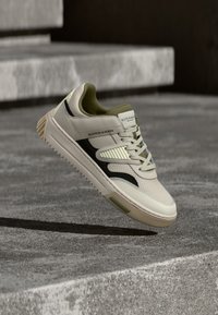 Adidași din piele crem cu accente verzi închise, panouri texturate, dungi negre pe lateral și o talpă robustă din cauciuc, ridicați deasupra unei suprafețe din beton.