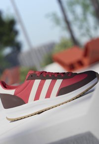 Roter und brauner Sneaker mit weißen Streifen und Gummisohle, im Freien auf einer weißen Oberfläche präsentiert.