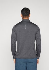 Pitkähihainen, tummanharmaa suorituskykyinen paita, jossa on teksturoitu viimeistely, puolivetoketjullinen kaulus ja brändäys keskellä selkää.