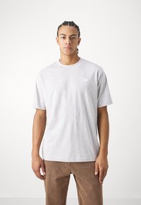 T-shirt en coton gris clair avec un col rond et des manches courtes. Présente un petit logo blanc sur la zone de la poitrine gauche.