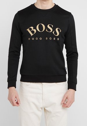 Sweat-shirt noir à manches longues avec logo doré "BOSS HUGO BOSS" porté avec un pantalon blanc, visible du cou à la taille.