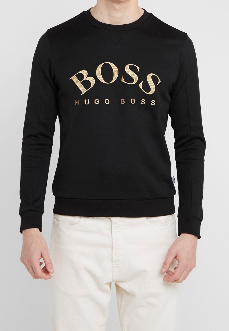 Sweat-shirt noir à manches longues avec logo doré "BOSS HUGO BOSS" porté avec un pantalon blanc, visible du cou à la taille.
