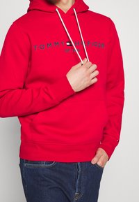 Sudadera roja con un bolsillo frontal, capucha ajustable con cordón y texto de logo azul "TOMMY HILFIGER EST. 1985." Tejido de mezcla de algodón. Corte casual.