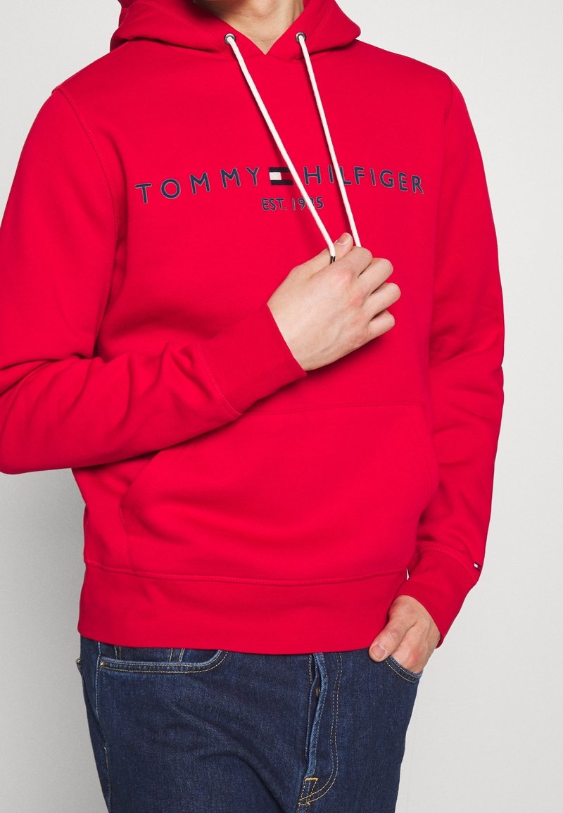 Sudadera roja con un bolsillo frontal, capucha ajustable con cordón y texto de logo azul "TOMMY HILFIGER EST. 1985." Tejido de mezcla de algodón. Corte casual.
