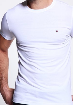 Mężczyzna ubrany w dopasowaną, gładką, białą koszulkę z krótkim rękawem, z małym prostokątnym logo Tommy Hilfiger umieszczonym na lewej piersi.