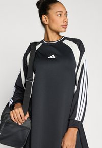 Juoda sportinė suknelė su baltais akcentų skydeliais ir Adidas logotipu. Turi ilgus rankovus, apvalią iškirptę ir laisvą siluetą. Laikoma juoda rankinė.