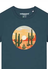 Teal katoenen T-shirt met een ronde afbeelding van een woestijnlandschap met cactussen, bergen en een zon, in aardetinten en zachte texturen.