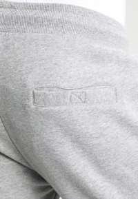Los pantalones de chándal grises presentan una cintura elástica y un pequeño detalle de parche cosido con un logo en la parte posterior, fabricados con una tela suave y texturizada.