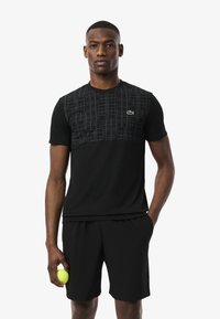 Uomo che indossa un completo sportivo nero con una maglia a fantasia, tiene in mano una pallina da tennis gialla e si presenta con un'espressione seria e sicura di sé.