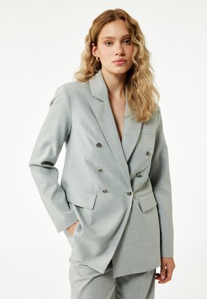 Junge Frau mit welligem blondem Haar, die einen hellgrauen, zweireihigen Anzug trägt, steht mit einer Hand in der Tasche vor einem einfarbigen Hintergrund.