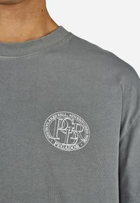 Pelēka kokvilnas T-krekla ar apaļu kakla izgriezumu, ar baltu apļveida logotipu, kurā ir teksts "SNIEGA KRASTI KRĪT, SEIKALAI PIEAUG, PEGADOR" uz priekšpuses.