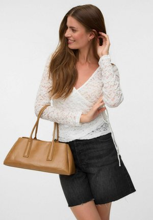 Femme aux longs cheveux bruns portant un haut en dentelle blanche et un short noir, tenant un sac à main en cuir beige et touchant ses cheveux.