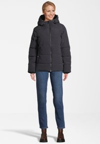 Gesteppte schwarze Winterjacke mit Kapuze, zentralem Reißverschluss, Druckknöpfen und gesticktem Muster. Kombiniert mit blauen Jeans und braunen Stiefeln.