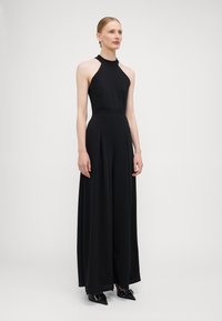 Black halter jumpsuit with a fitted bodice, wide pleated legs, and smooth fabric. Notable pointed heels with decorative hardware.