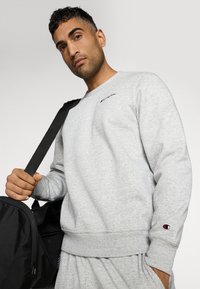 Gray sweatshirt with ribbed cuffs and waistband, featuring a small black logo on the chest. Model holding a black duffle bag.
