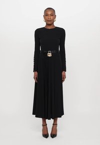 Must mustael dress, millelõustega, ent kitsenev ülemine osa ja voolav seelik. Varustatud musta vöödiga, millel on metallist ruudukujuline pandla. Kannab musti kontsasid.