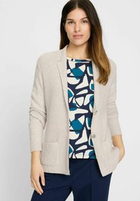 Hellgraue Strickjacke mit geripptem Muster, Ein-Knopf-Verschluss und zwei vorderen Taschen, darüber getragen ein Oberteil mit geometrischem Muster.