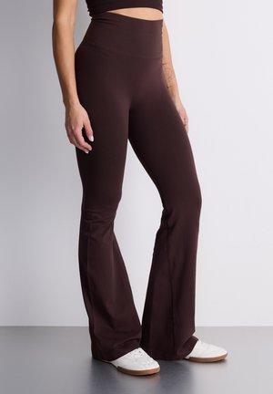 Leggings marroni a zampa d'elefante in tessuto elasticizzato con vita alta. Indossati con scarpe da ginnastica bianche, evidenziando una texture liscia e linee pulite.