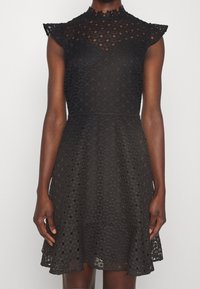 Svart spetsklänning med korta ärmar, hög krage och en utsvängd kjol med ett hexagonmönster. Strukturerad tyg med ett fodrat inre.